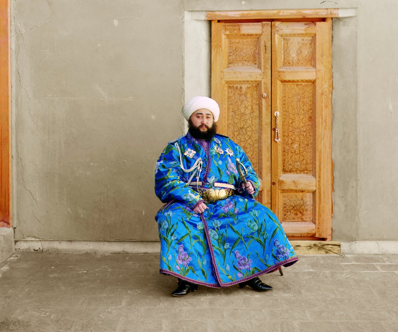 Цветная фотография Прокудина-Горского: эмир Бухары Алим-хан в синем расшитом халате и белой чалме сидит у резной деревянной двери